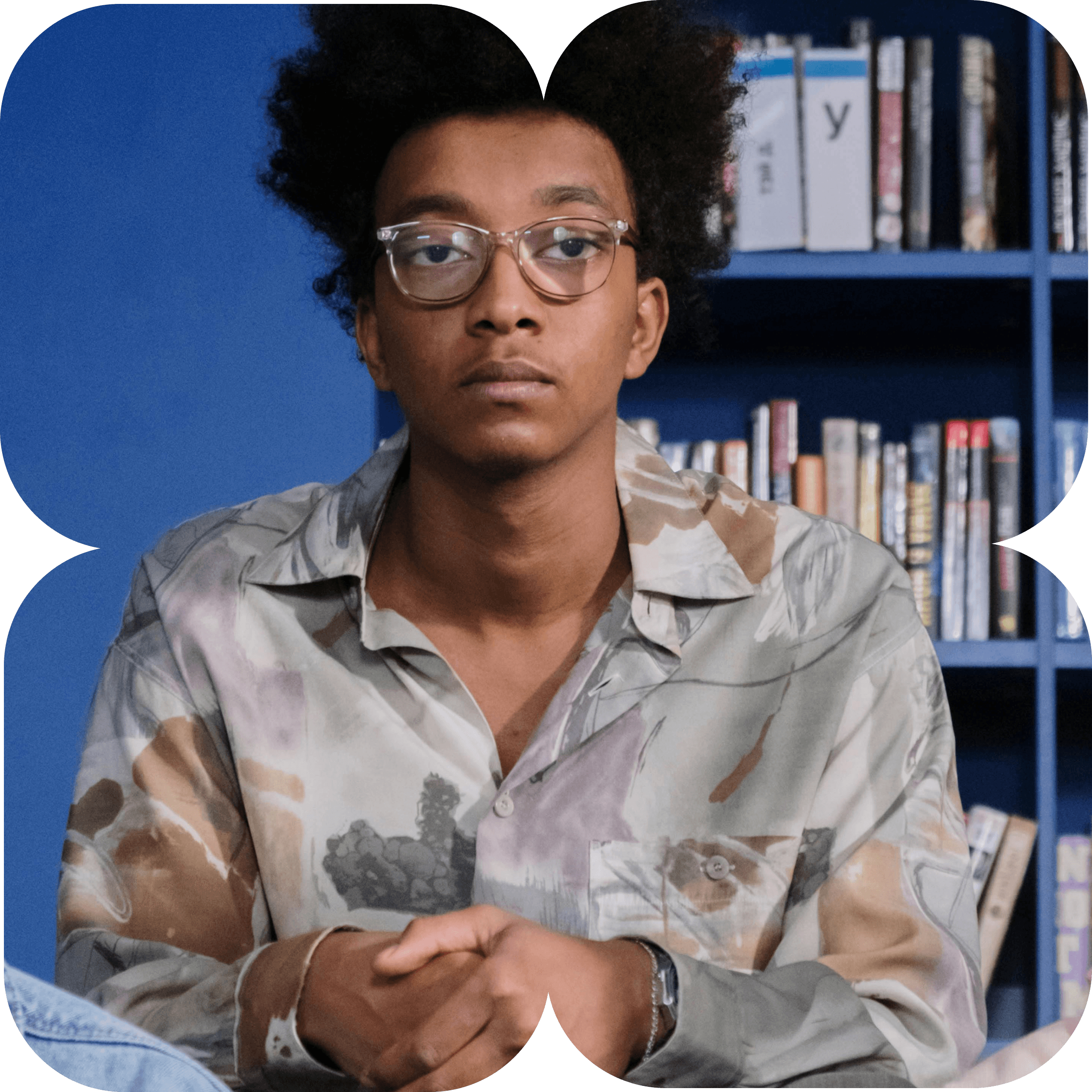 Un joven con gafas y cabello rizado sentado frente a una estantería con libros, con una expresión seria.